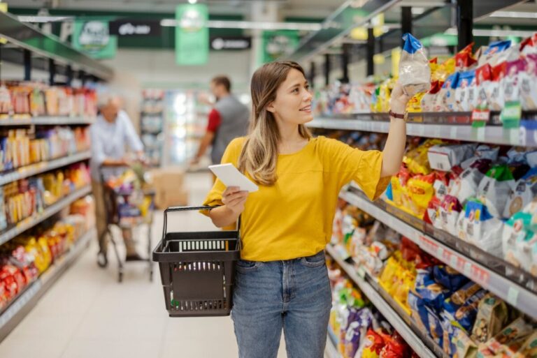 Woman in yellow top and jeans holding grocery basket and shopping for CPG products