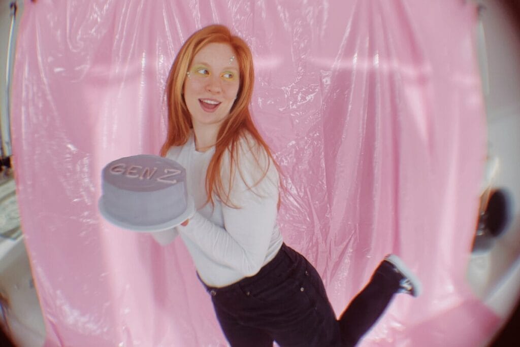 Teenage girl with red hair, white shirt and black pants holding light purple cake that says Gen Z