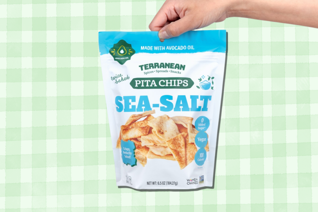 Hand holding bag of Terranean Avocado Oil Pita Chips over green gingham background